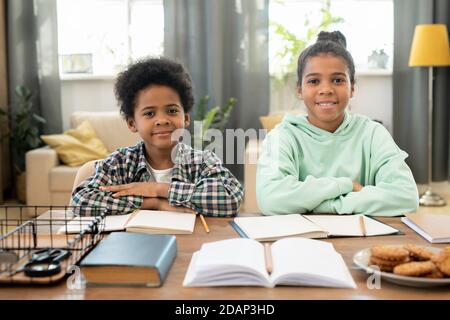 Felice ragazza elementare e ragazzo in casualwear seduto al tavolo in soggiorno Foto Stock