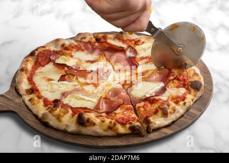 Pizza con pancetta o speck e formaggio brie e souce di pomodoro. Prodotto italiano su tagliere in legno con ruota a coltello su sfondo marmoreo Foto Stock