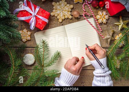 Le mani della donna scrivono la lettera di Natale, wishlist o fare la lista su carta del notepad su sfondo di legno con la decorazione di Natale, la vista superiore Flatlay copia spac Foto Stock