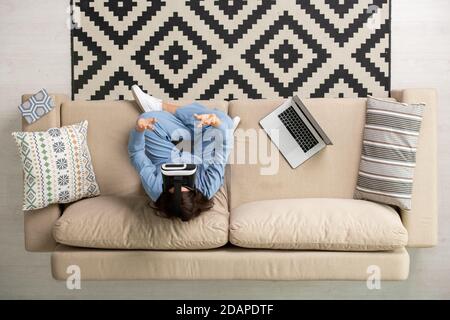 Panoramica di giovane donna brunetta in pigiama blu e vr cuffie che funzionano sul divano Foto Stock