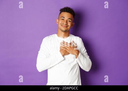 Ritratto di felice sorridente afroamericano, sentendosi grato e toccato con bel gesto, tenendo le mani sul cuore, ringraziando per qualcosa Foto Stock