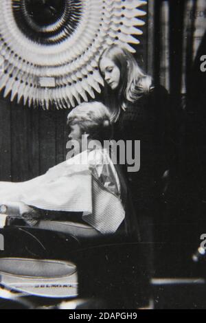 Bella fotografia in bianco e nero d'epoca degli anni '70 di una giovane donna che dà un taglio di capelli a un ragazzo. Foto Stock