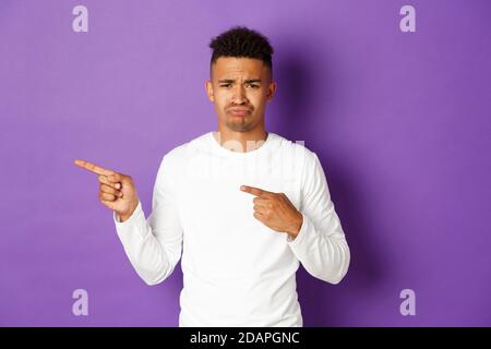 Ritratto di triste e deluso giovane afro-americano, lamentando e sulking, puntando le dita lasciate a qualcosa di cattivo, in piedi su viola