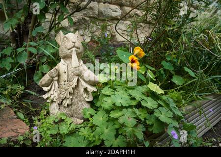 Cast statua giardino in cemento del personaggio Peter Rabbit mangiare una carota in un giardino troppo cresciuto bordo contro una pietra parete Foto Stock