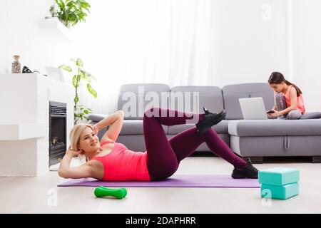 La donna sta facendo yoga online con il laptop durante l'isolamento di sé nel suo soggiorno, senza attrezzi di allenamento, suggerimenti per la meditazione per i principianti. Sua figlia è Foto Stock