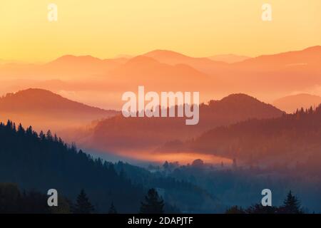 Paesaggio con belle montagne, campi e foreste ricoperti di fitta nebbia mattutina. Un'alba interessante illumina i dintorni. Fantastico autunno d Foto Stock