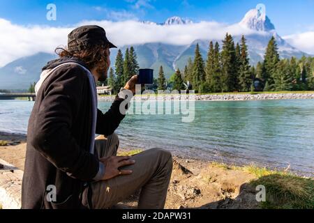 Vista laterale foto di un maschio di mezza età seduto su una panchina, con un caffè mattutino in mano mentre si gode la vista di un fiume cristallino e le montagne Foto Stock