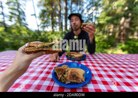 Fuoco selettivo di due persone adulte che hanno una casa deliziosa pasto fatto di panini ad un tavolo da picnic all'interno di un parco nazionale esterno Foto Stock