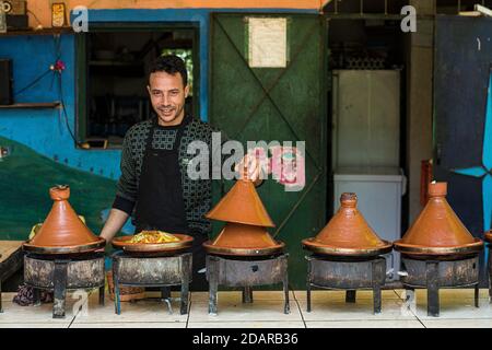 Tajine, un uomo offre cibo tradizionale marocchino preparato su fuoco di carbone in pentole Tajine tipiche in uno stand Foto Stock