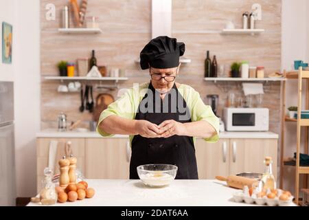 Spezzare le uova in farina di grano seguendo la ricetta tradizionale in cucina casalinga. Cuoco di pasta anziano che coglie l'uovo sulla ciotola di vetro per la ricetta della torta in cucina, mescolando a mano, impastando. Foto Stock