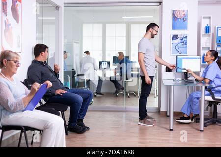 Giovane uomo che parla con l'infermiera alla reception.elevator dell'ospedale. Paziente in attesa di essere invitato nella sala esame. Donna anziana che compila documenti. Foto Stock