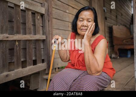 Tailandese anziana donna in collo rotondo senza maniche con mal di testa e. preoccupato volto stressato in vecchia casa di legno Foto Stock