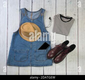 Still life di abiti, gonna in denim con ungarees, camicia a righe, stivali militari, cappello, smartphone e cuffie, su uno sfondo di legno bianco Foto Stock