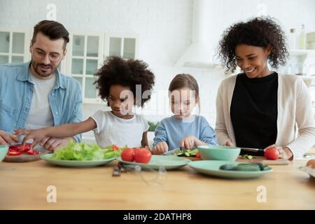 Felice famiglia multirazziale con i bambini che cucinano l'insalata, tagliando insieme le verdure Foto Stock