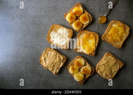 Colazione dolce foto vista dall'alto. Tostature con condimenti diversi: Burro di arachidi, marmellata, burro e banana caramellata. Sfondo grigio scuro con texture Foto Stock