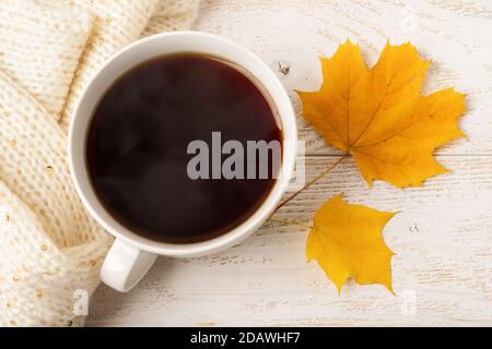 Grande tazza bianca di caffè fumante caldo e due foglie di caduta gialle vicino a sciarpa di maglia beige sopra la superficie rustica di legno bianco. Accogliente concetto autunnale. Foto Stock