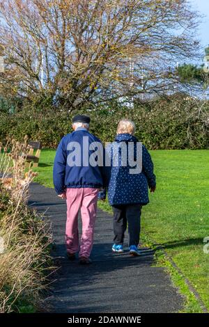 Una coppia anziana che cammina in un parco in un giorno d'autunno luminoso e soleggiato l'uomo indossa pantaloni color salmone e un berretto bretone. Foto Stock