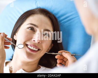 Donna bruna sorridente esaminata dal dentista in clinica dentale. Mani di un medico che tiene gli strumenti dentali vicino alla bocca del paziente. Denti sani AN Foto Stock