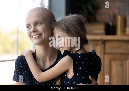 Giovane madre senza peli in piedi dalla finestra abbracciando la piccola figlia Foto Stock