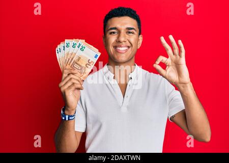 Giovane arabo che tiene 50 banconote in euro facendo ok segno con le dita, sorridente amichevole gesturing eccellente simbolo Foto Stock