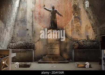 Statua del prete napoletano Gaetano Barbati circondata da ossa umane impilate all'interno del Cimitero delle Fontanelle (Cimitero delle Fontanelle) a Napoli, Campania, Italia. L'ex caserma (ossario) situata in una grotta del distretto di Materdei era il luogo in cui si sviluppò a Napoli il culto spontaneo della devozione ai resti di morti senza nome. I difensori del culto hanno pagato visite ai teschi senza nome nell'ossario per adottarli e persino dare i nomi dei teschi. I teschi adottati furono collocati all'interno della scatola decorata e diventarono oggetto di regolare offerta orante e votiva. Foto Stock