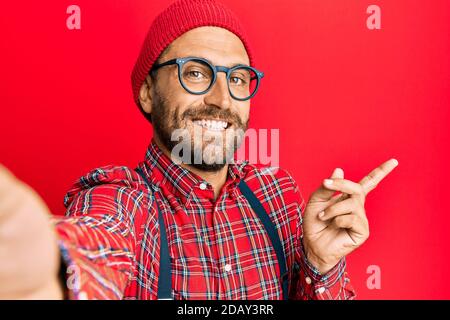 Bell'uomo hipster con barba scattando una foto selfie con smartphone sorridente che punta con mano e dito verso laterale Foto Stock