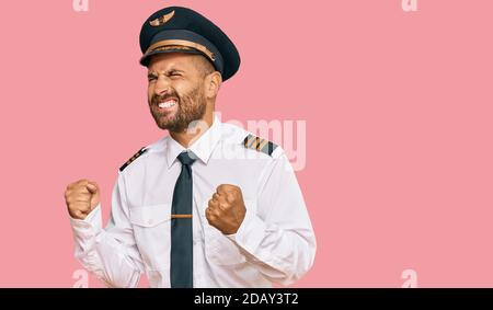 Bell'uomo con barba che indossa l'uniforme pilota di aeroplano eccitato per il successo con le braccia sollevate e gli occhi chiusi celebrando la vittoria sorridente. Vincitore concep Foto Stock