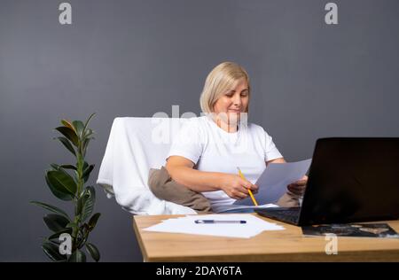 Lavoro online senior. Donna è seduta in poltrona a casa e lavora con un computer. Concetto online senior e concetto pandemico. Foto Stock