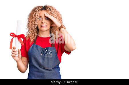 Giovane donna bionda con capelli ricci che indossa grembiule da parrucchiere e che tiene diploma stressato e frustrato con la mano sulla testa, sorpreso e arrabbiato viso Foto Stock