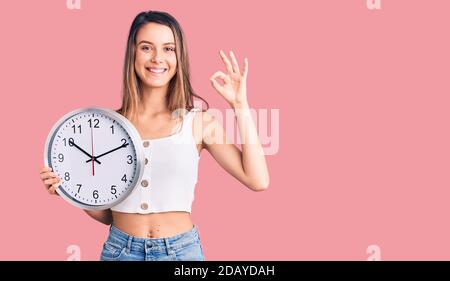 Giovane bella ragazza che tiene grande orologio facendo ok segno con le dita, sorridente amichevole gesturing eccellente simbolo Foto Stock