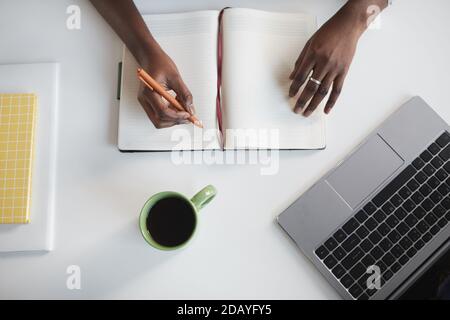 Vista dall'alto minima di una donna afroamericana irriconoscibile che scrive nel pianificatore sul posto di lavoro di un'azienda bianca Foto Stock