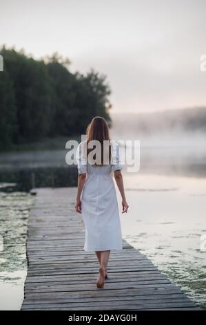 Vista posteriore di giovane donna in abito bianco in piedi da sola sul ponte pedonale e fissando al lago. Mattina fredda con una nebbia sull'acqua. Foto Stock