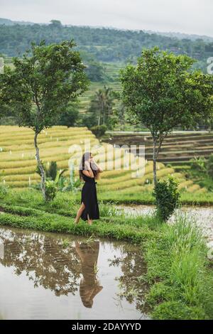 Donna che cammina contro le fantastiche terrazze di riso di Jatiluwih a Bali. Bellezza e armonia nella natura. Indossabile in un lungo abito nero. Foto Stock