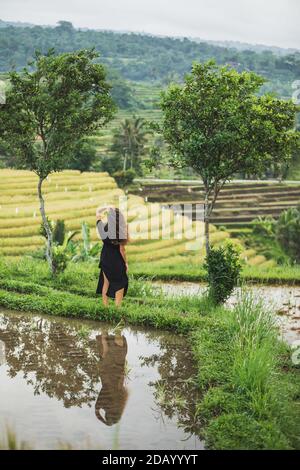 Donna che cammina contro le fantastiche terrazze di riso di Jatiluwih a Bali. Bellezza e armonia nella natura. Indossabile in un lungo abito nero. Foto Stock