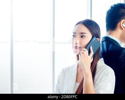 Coppia asiatica d'affari che parla con il telefono cellulare. Giovane bella donna fiduciosa che chiama con il telefono cellulare e in piedi con uomo d'affari in tuta su h. Foto Stock