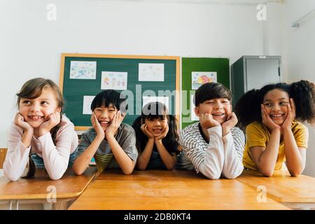 Amici allegri della scuola multietnica di fronte a fotocamera con mani sul mento, felicità, togetherness, amicizia. Ritratto dei bambini che sorridono in classe Foto Stock