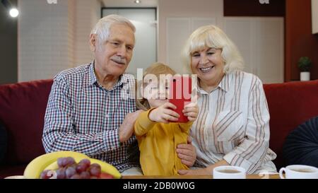 Coppia senior nonni con bambina nipotina passare il tempo a casa insieme, seduto sul divano in soggiorno, utilizzando il telefono cellulare. Giocare, fare foto selfie insieme sullo smartphone Foto Stock