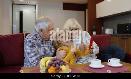 Coppia senior nonni con bambina nipotina passare il tempo a casa insieme, seduto sul divano in soggiorno, utilizzando il telefono cellulare. Giocare, fare foto selfie insieme sullo smartphone Foto Stock