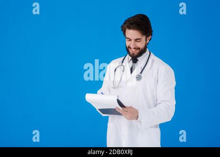 un giovane medico con la bearded in un cappotto medico bianco sopra uno sfondo blu scrive i reclami del paziente su un tablet con documenti e medici Foto Stock