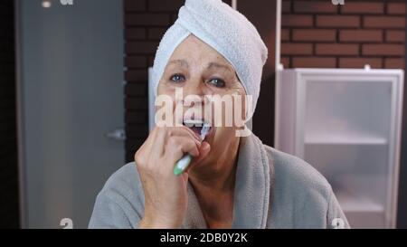 Vecchia nonna anziana con asciugamano in accappatoio spazzolando i denti e guardando in uno specchio. Nonna anziana che fa igiene del mattino dopo la doccia in bagno di lusso a casa. Punto di vista. Shot POV Foto Stock