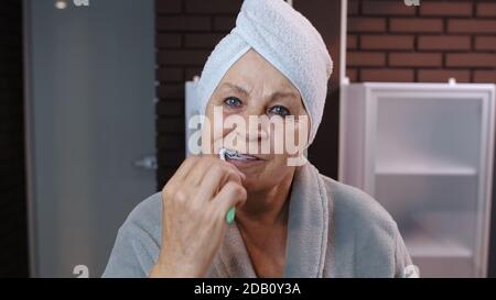 Bella nonna anziana con asciugamano spazzolando i denti e guardando in uno specchio. Nonna anziana che fa igiene del mattino dopo la doccia in bagno di lusso a casa. Punto di vista. Shot POV Foto Stock
