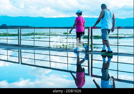 Coppia asiatica matura esercizio sulla terrazza vicino al lago dopo la pioggia, montagna blu e nuvoloso in background. Phayao, Thailandia. Foto Stock