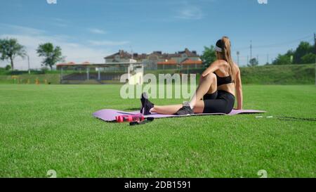 Irriconoscibile ragazza sottile che indossa abbigliamento sportivo in estate soleggiato giorno. Vista laterale della giovane donna che allunga il corpo sul tappetino viola dopo la corsa allo stadio. Flessibilità, concetto di stretching. Foto Stock