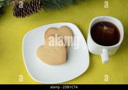 Due biscotti a forma di cuore su un piatto bianco con una tazza di tè su sfondo giallo con un ramo di un albero di Natale artificiale con un cono. Selectiv Foto Stock