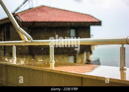 Ringhiera metallica sul balcone e gocce di pioggia che cadono dal cielo sono rotte sul parapetto, tetto di tegole rosse della casa. Visualizza dalla finestra. Foto Stock