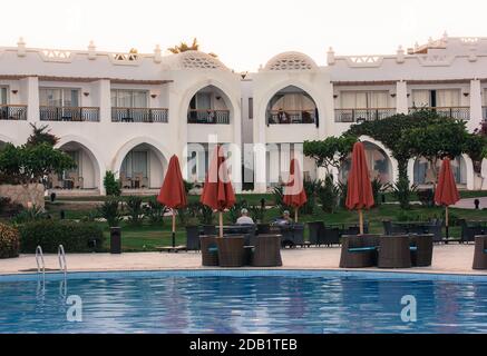 Area lounge vicino alla piscina d'acqua blu dell'hotel in Egitto. Tavoli e sedie per il relax dei turisti, blocchi bianchi, ombrelli rossi Foto Stock