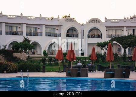 Area lounge vicino alla piscina d'acqua blu dell'hotel in Egitto. Tavoli e sedie per il relax dei turisti, blocchi bianchi, ombrelli rossi Foto Stock