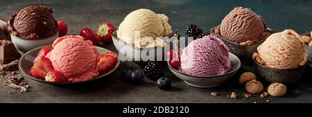 Ciotole di gelato artigianale con ingredienti freschi tra cui macaroons, varietà di bacche, baccelli di vaniglia e cioccolato in un banner panorama scuro Foto Stock