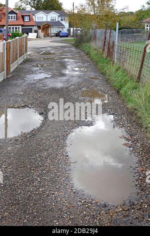 Scena di strada non adottata unmade unsopramed puddled & POT holed trascurato strada bagnata dopo la pioggia invernale al di fuori delle case del villaggio e fattoria Field Essex Inghilterra Regno Unito Foto Stock