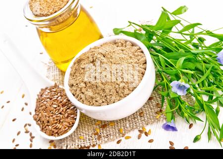 Farina di semi di lino in una ciotola, semi di lino in un cucchiaio, olio in vaso di vetro su un tovagliolo di burlap, foglie e fiori di lino su sfondo di legno Foto Stock